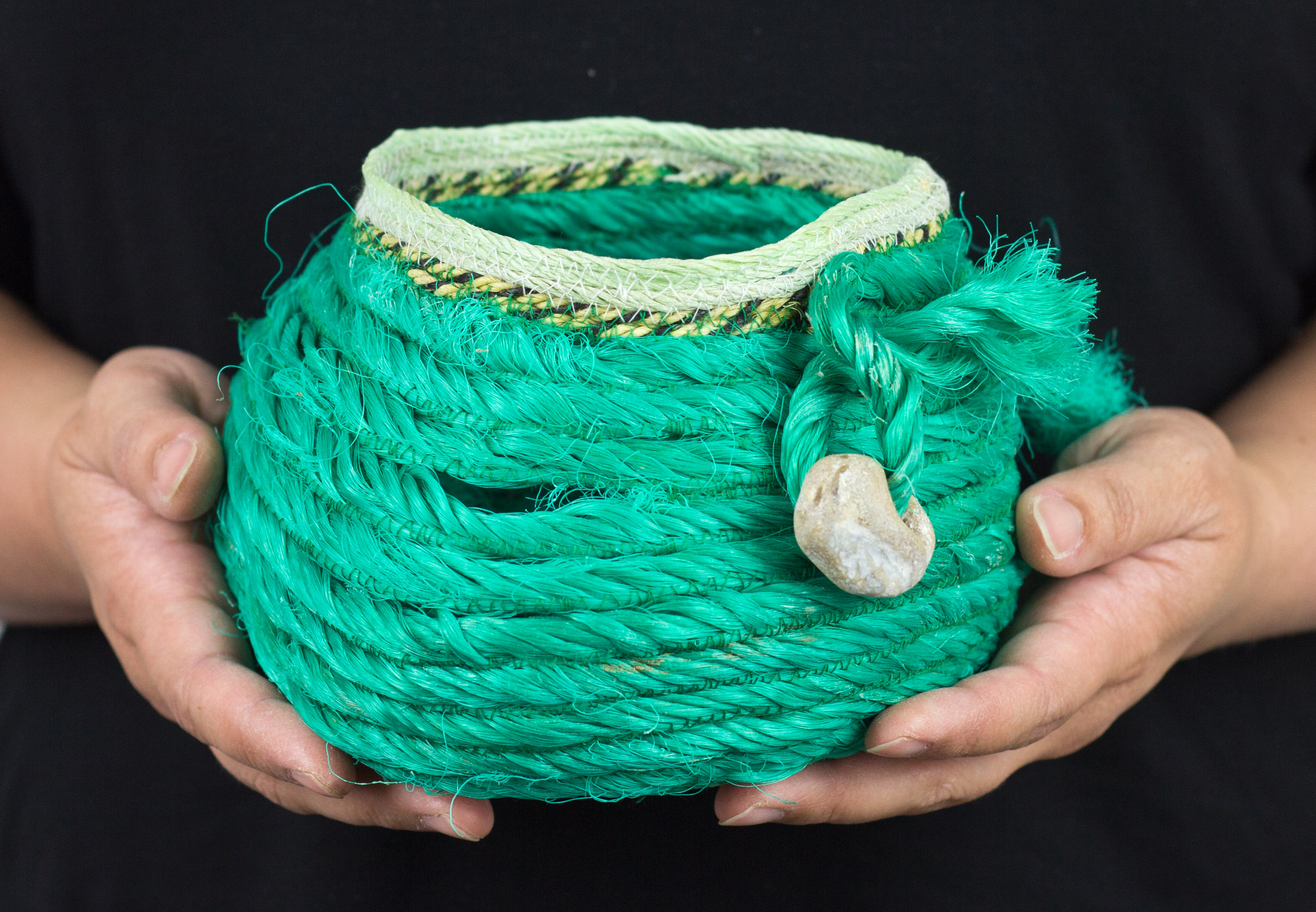 hands holding a basket made of crab rope, the rope is turquoise and black with a stone from the beach hanging from it.