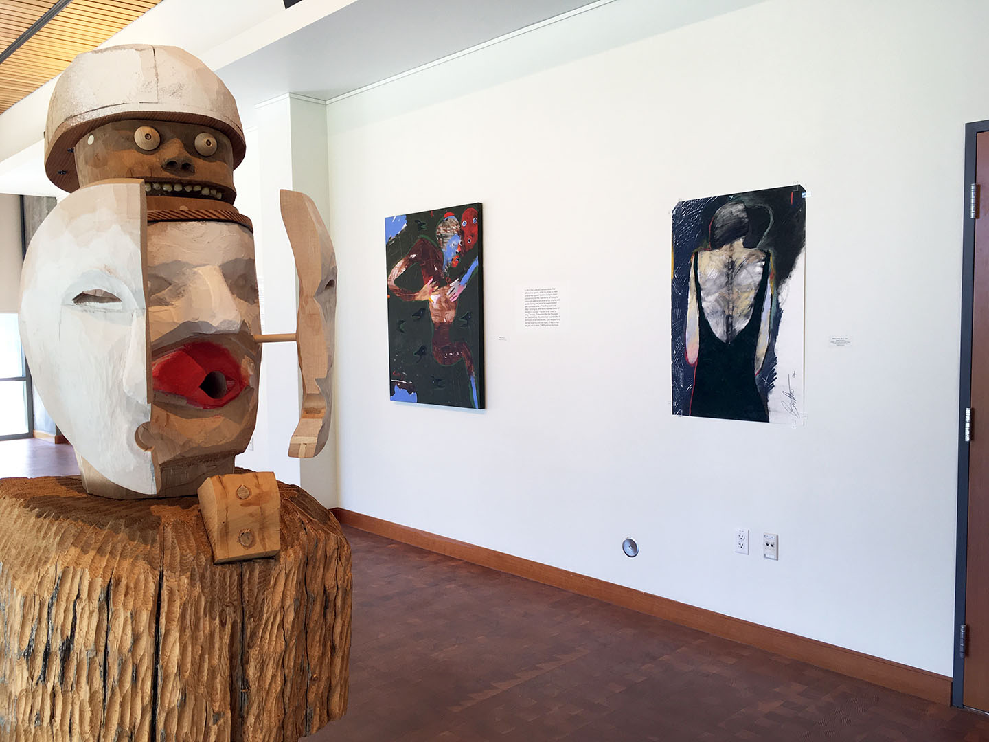view of wooden sculpture in foreground with two faces opening to view a secondary face below with red lips and white face. white gallery walls behind with 2d artwork on them.