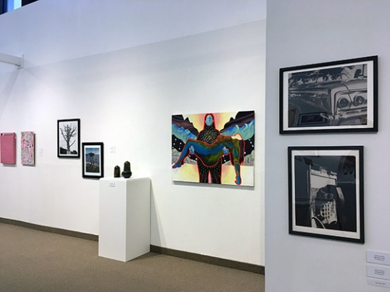view of gallery with white walls, and two photographs in the foreground and a colorful painting 